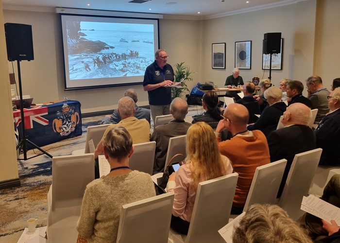 Jim Kerr presenting his Secretary's reports during the AGM section of the Gathering.
      The background photograph is one taken on Tristan in 1985/86 by the late Bob Kauders. Photo: Peter Millington